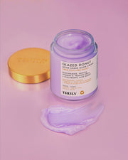 A jar of Truly Beauty’s Glazed Donut After Shave Glow Cream with an open gold lid rests on a pink surface, with some light purple cream smoothly spread in front of it.