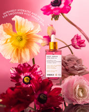 A bottle of Truly Beautys Soft Serve After Shave Duo with a dropper rests amid colorful flowers on a pink background. White text above says: Hydrating and ingrown-fighting for smoother, intensely healed post-shave skin.