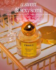 The Vanilla Baby Fragrance Duo by Truly Beauty, featuring a sweet & sexy vanilla scent with notes of amber, pistachio, and caramel, rests on a gold tray beside luxe body oil and blurred pink flowers in the background.