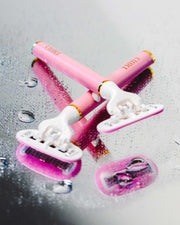 Two razors from Truly Beauty’s The Smooth Legend - Complete Collection lie crossed on a wet, reflective surface with scattered water droplets—perfect for elevating your shaving routine.