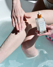 While soaking in a bathtub, someone applies Truly Beautys Coco Cloud Shave Routine from a glass bottle with a dropper onto their leg as part of their luxury shaving ritual.