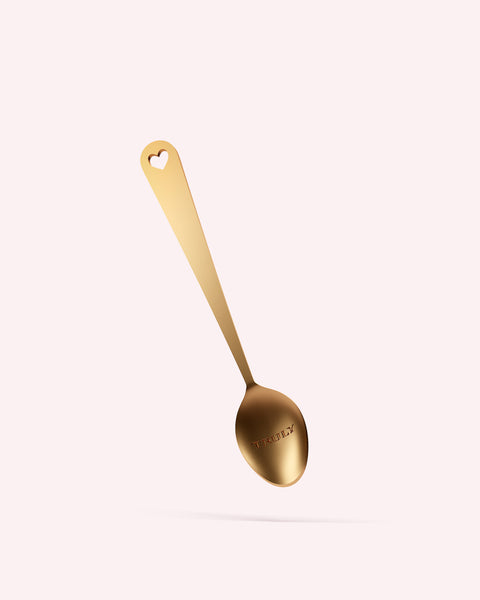 The Truly Beauty Mini Gold Spoon features a heart-shaped cutout in the handle and engraved text. Shown on a light pink background, its the perfect product scoop for skincare application.