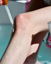 A person applies Truly Beauty’s Coco Cloud Shave Routine post-shave oil to their bare leg with a dropper, glistening droplets on the skin and a pink towel in the background.