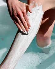 While relaxing in a bubble bath, someone smooths a thick layer of Truly Beauty’s Coco Cloud Shave Routine onto their leg as soapy bubbles float on the water around them.