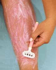 A person uses the Truly Beauty Razor Cartridges with a pink-handled razor and shaving cream to shave their leg over a pale blue surface—ideal for silky smooth results in the bath or shower.