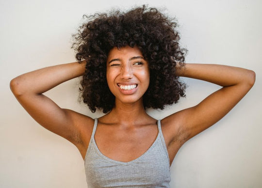 should you moisturize before shaving | A woman sits outdoors in sunlight, pouring Truly Beauty’s Sun Drops Bronze Body Glow from a brown bottle into her palm. Only her torso and hands are shown, emphasizing her radiant, bronzed skin