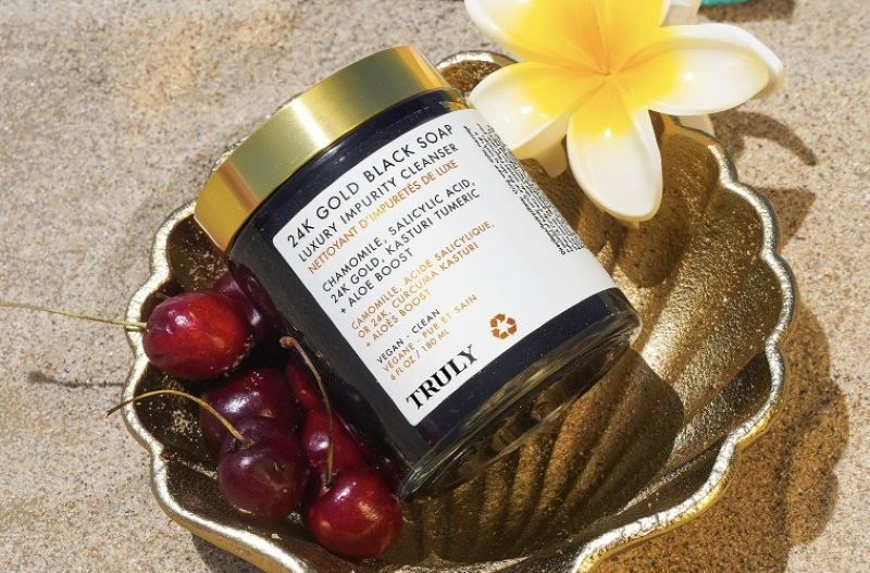 advantages of african black soap | Gold shell shaped bowl featuring jar of Truly's 24k gold black soap cleanseer and cherries on sandy beach