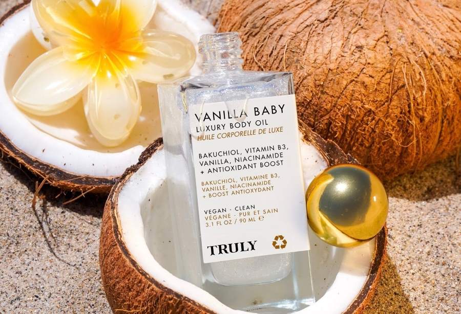 bakuchiol benefits for skin | Open bottle of Truly's Vanilla Baby Luxury Body Oil sits in coconut shell on sandy background