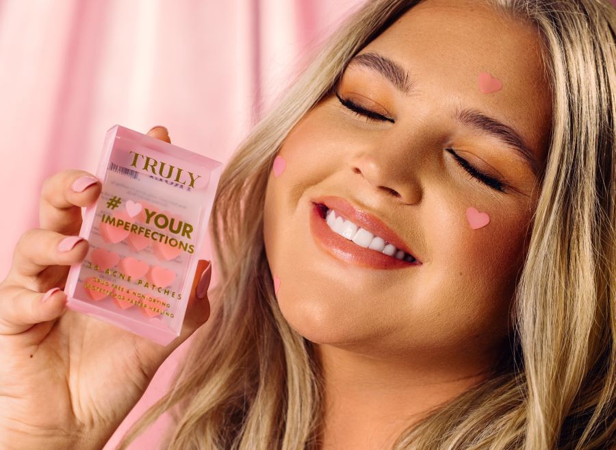 Woman with pink heart pimple patches on face holds a pack of Truly heart pimple patches 