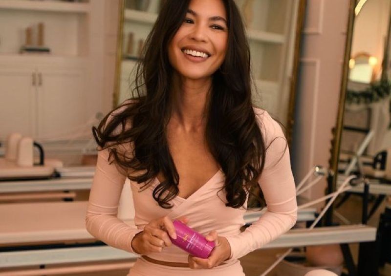 best ingredients for dark spots | A smiling woman with long, dark wavy hair wears a light pink long-sleeve wrap top while holding Truly's Unicorn Fruit deodorant, set against a softly lit background featuring fitness equipment.