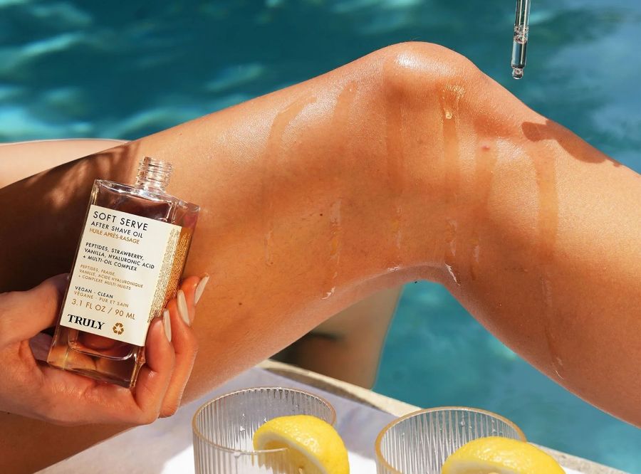body oil vs lotion | A person's oiled leg next to a pool, holding a bottle of Truly Soft Serve After Shave Oil with lemon drinks in the foreground.