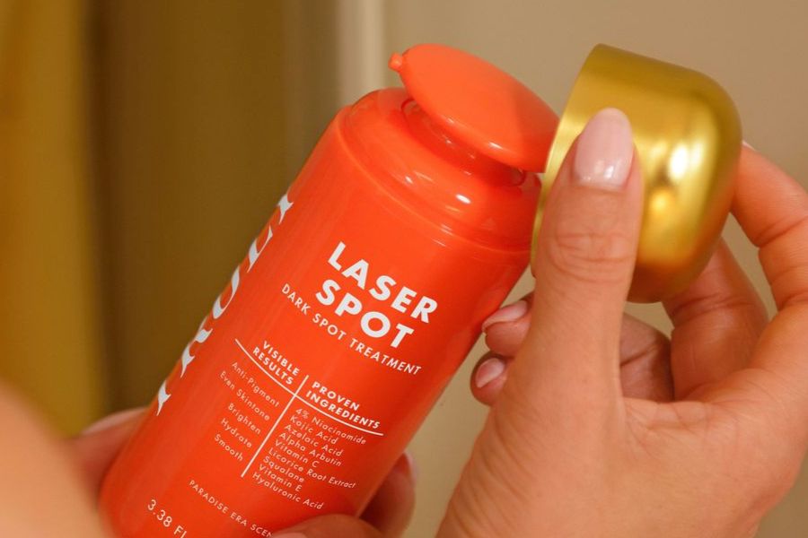 skincare routine dark spots | Close-up of a person's hands holding a bright orange bottle of Truly Dark Spot Treatment, with the gold lid lifted off.