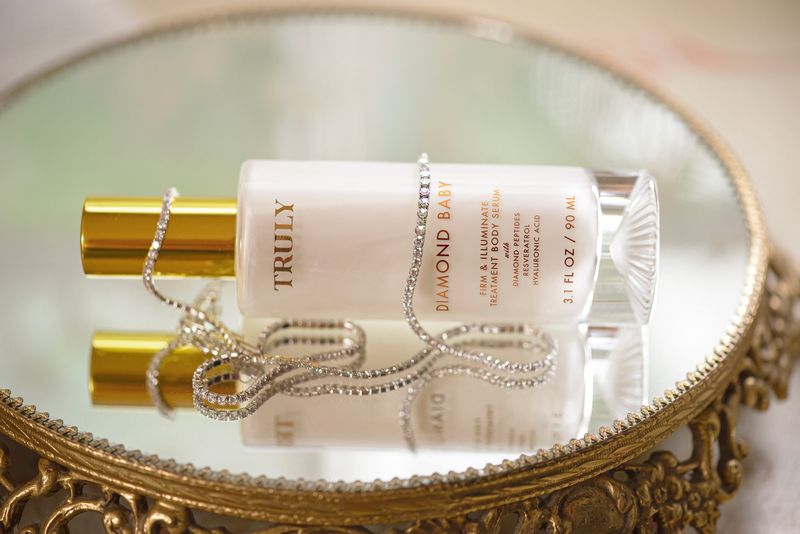 Truly's Diamond Baby Treatment Body Serum with diamond peptides wrapped in a diamond necklace, placed sideways on gold mirror