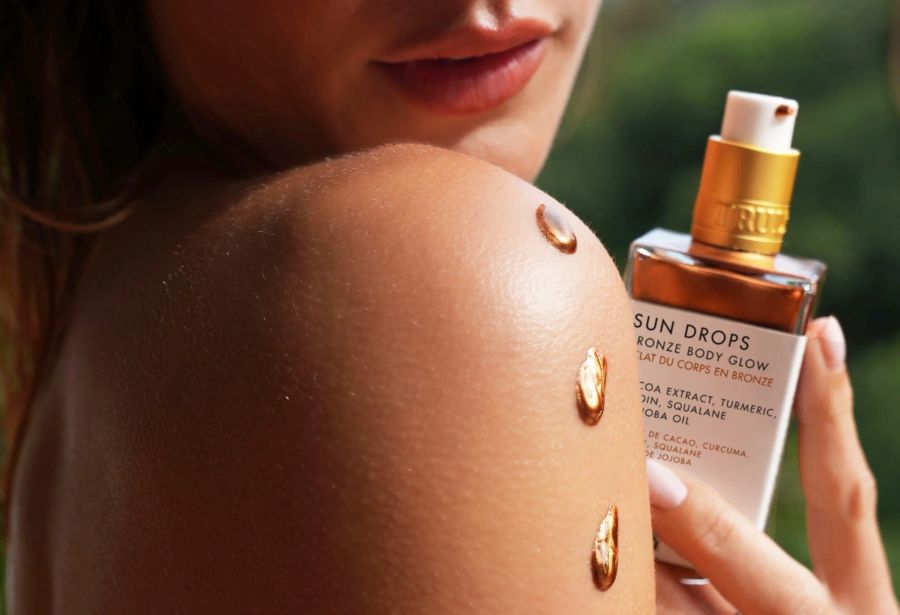 how long does self tan last | A woman holds a bottle of Truly Beauty Sun Drops Bronze Body Glow, displaying glowing drops on her shoulder against a blurred outdoor background.