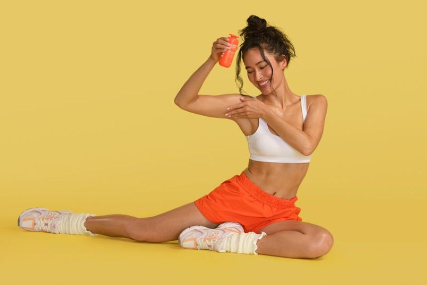 Woman wearing white sports bra and orange shorts applies body hyperpigmentation serum to underarms 