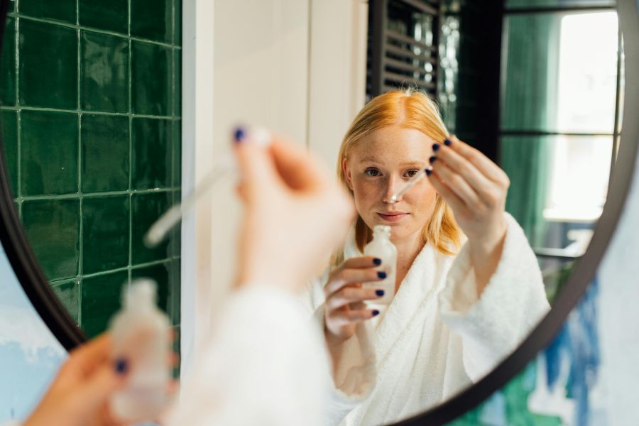 serum vs moisturizer | Woman in front of bathroom mirror wearing white bathrobe and holding a bottle and a dropper