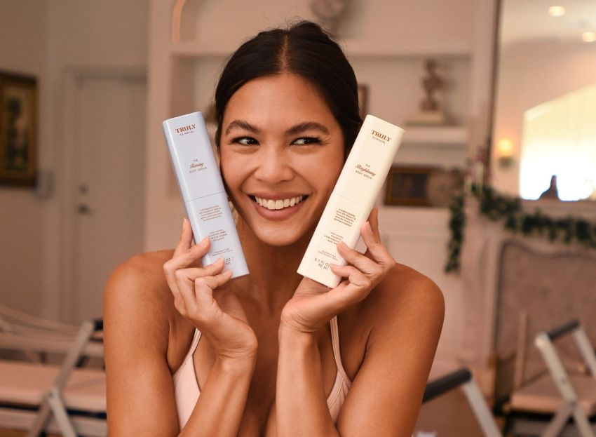 A woman with a bright smile posing with two Truly Beauty skincare products; one bottle is light blue - The Firming Body Serum - and the other is white - The Brightening Body Serum | spring skincare tips
