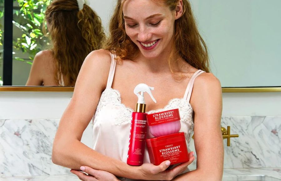 A woman in a white satin top holds three red Strawberry Blossoms skincare products for uneven skin texture in a bathroom setting.