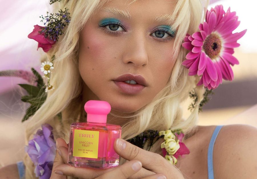 blonde woman with pink flowers in hair holds bottle of Truly Unicorn fruit perfume