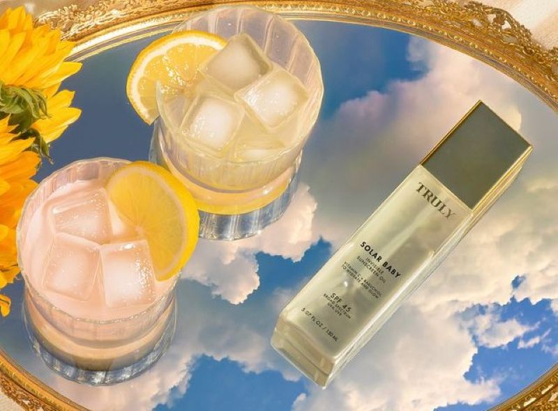 A bottle of Truly Solar Baby invisible sunscreen oil next to two iced drinks with lemon slices on a mirror reflecting a cloudy blue sky.