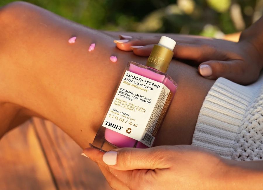 A person holds a bottle of Truly Smooth Legend After Shave Serum next to three small pink dots on their leg, showcasing one of the best products for KP for smoothing skin texture.