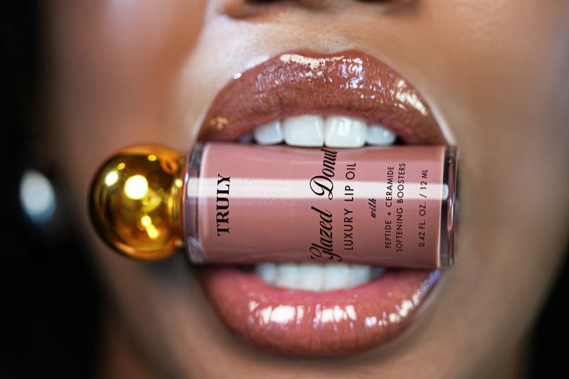 Close-up of glossy lips holding a Truly Beauty Glazed Donut Lip Oil with a golden cap between the teeth. | What's the best lip plumper 2025