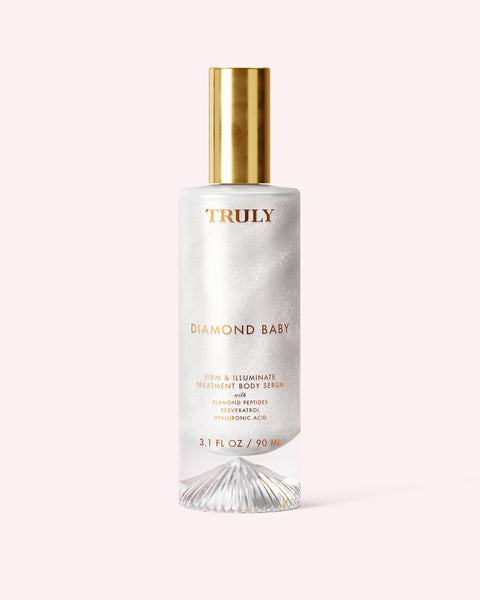 A bottle of Truly Beauty Diamond Baby Treatment Body Serum with a gold cap, clear glass, and iridescent white liquid features diamond peptides for glowing skin—displayed against a pale pink background.