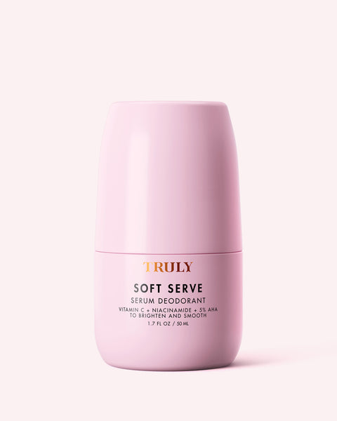 A pink cylindrical container of Truly Beauty Soft Serve Serum Deodorant, featuring gold and black text, describes it as a vitamin C deodorant with niacinamide & 5% AHA to brighten and smooth. Size: 1.7 fl oz (50 ml) on a light pink background.