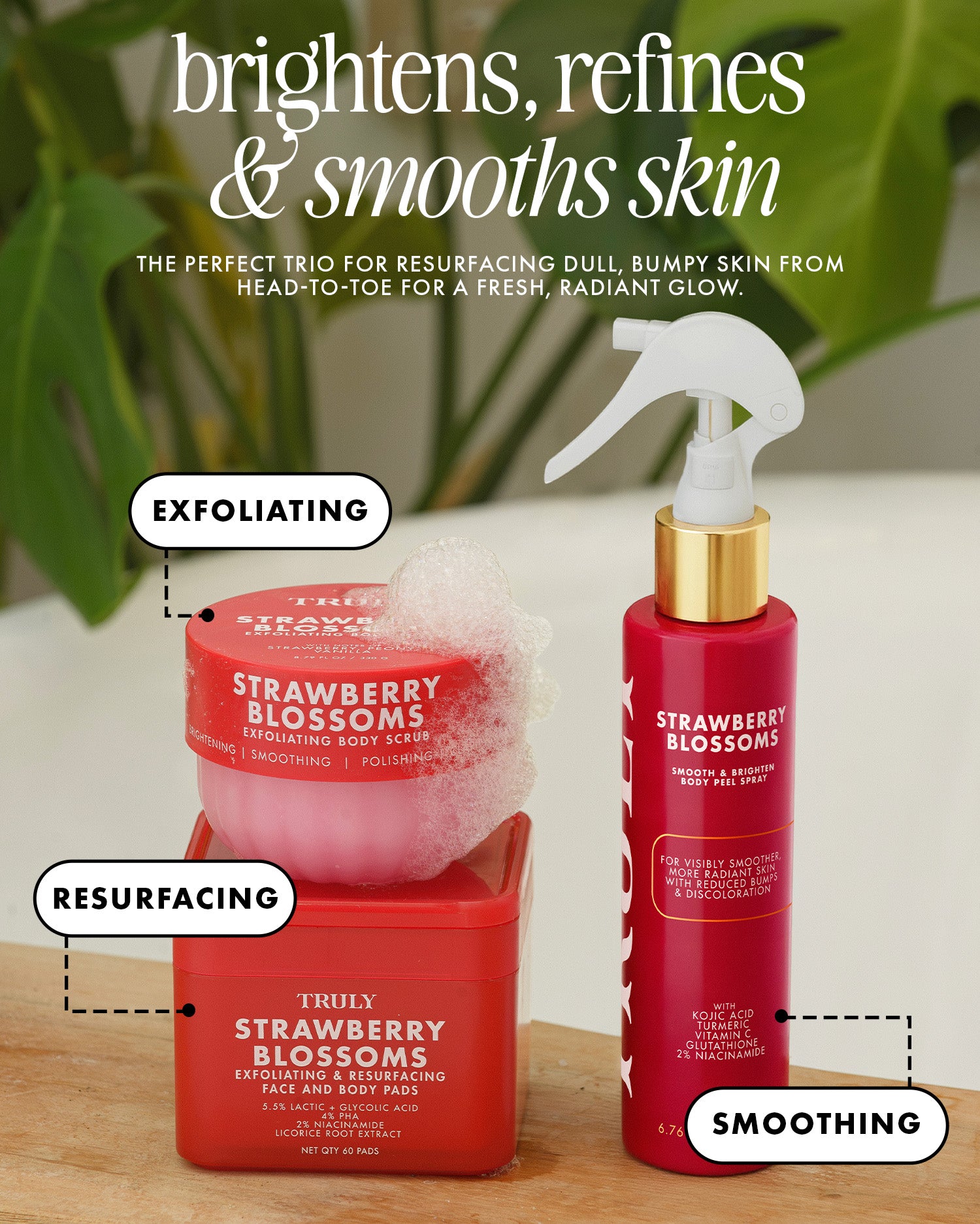 Three red skincare products from Truly Strawberry Blossoms are arranged on a wooden surface with green leaves in the background. The set includes an exfoliating scrub in a square jar with a white sponge on top, a resurfacing acid treatment in a rectangular jar, and a smoothing spray in a cylindrical bottle with a gold collar and white sprayer.
