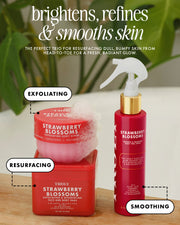 Three red skincare products from Truly Strawberry Blossoms are arranged on a wooden surface with green leaves in the background. The set includes an exfoliating scrub in a square jar with a white sponge on top, a resurfacing acid treatment in a rectangular jar, and a smoothing spray in a cylindrical bottle with a gold collar and white sprayer.