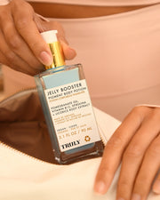 A person holds a bottle of Jelly Booster Pigment Body Potion by Truly Beauty above an open cream-colored bag. The label highlights vitamin B12 and natural ingredients known for hyperpigmentation serum benefits.
