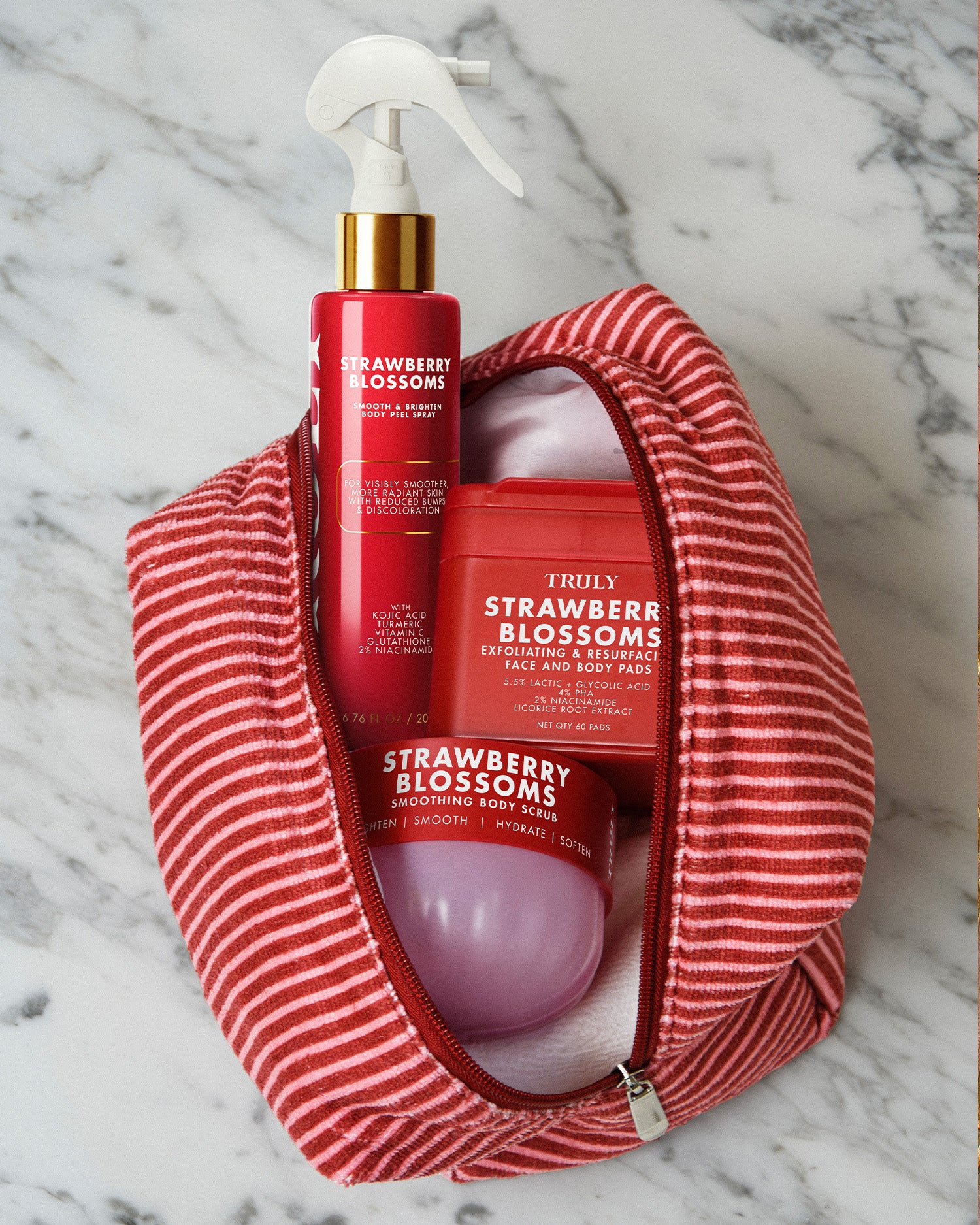 The Truly Beauty Strawberry Blossoms Complete Collection—featuring a spray, body mask jar, and round container in elegant red and pink hues—peeks out from a partially unzipped red and white striped toiletry bag set on a marble surface. Designed to enhance your skin cycling routine, this collection is ideal for addressing concerns like keratosis pilaris and helps you achieve smoother, strawberry skin.
