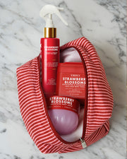 The Truly Beauty Strawberry Blossoms Complete Collection—featuring a spray, body mask jar, and round container in elegant red and pink hues—peeks out from a partially unzipped red and white striped toiletry bag set on a marble surface. Designed to enhance your skin cycling routine, this collection is ideal for addressing concerns like keratosis pilaris and helps you achieve smoother, strawberry skin.
