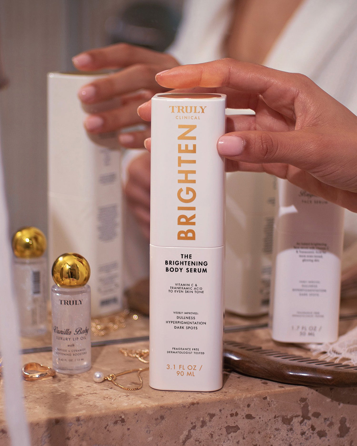 A person's hands hold a white bottle of Truly Clinical Brighten The Brightening Body Serum on a beige stone countertop with other skincare products in the background.