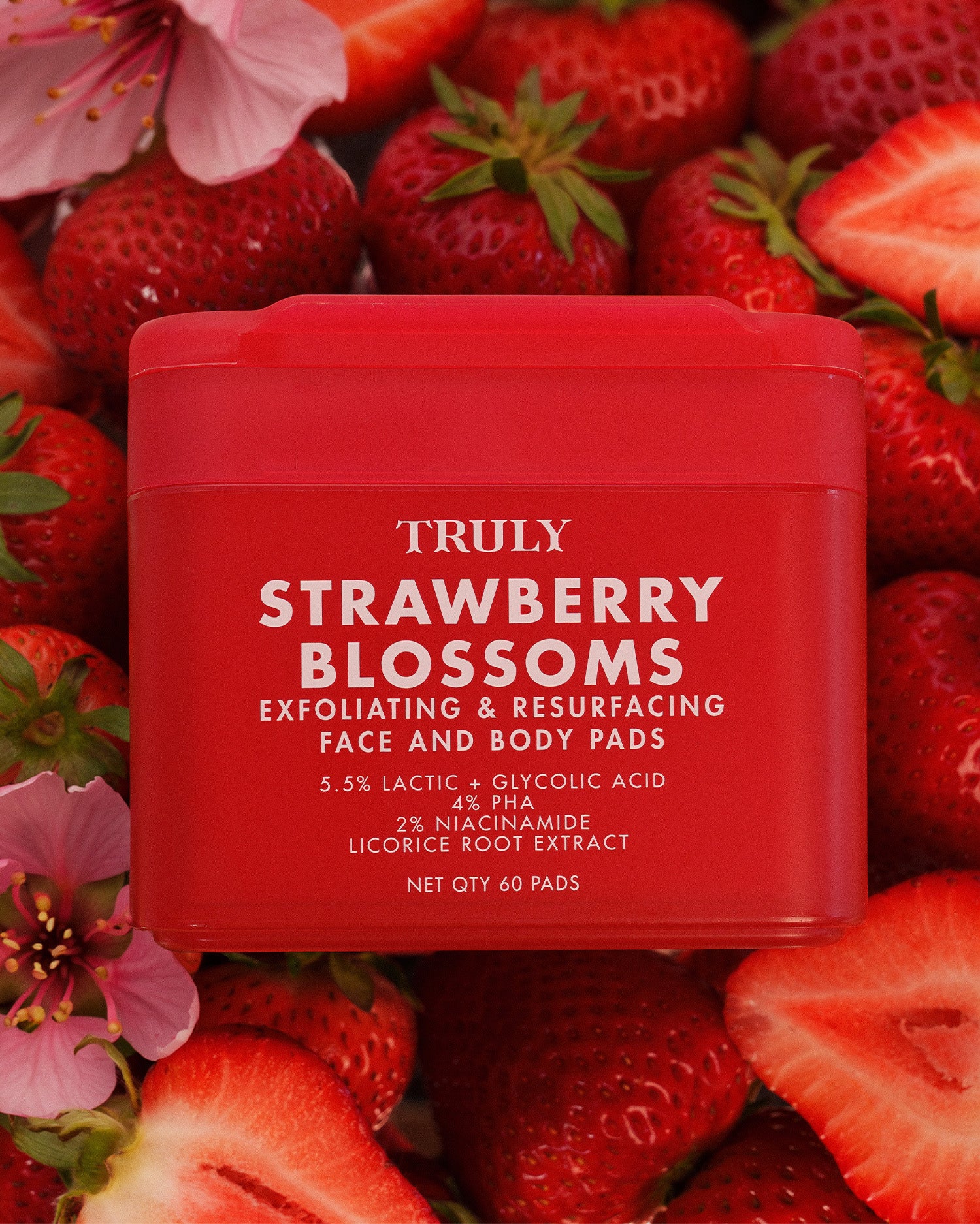 A red tub of Truly Strawberry Blossoms exfoliating and resurfacing face and body pads, surrounded by fresh strawberries and pink blossoms.