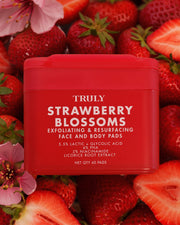 A red tub of Truly Strawberry Blossoms exfoliating and resurfacing face and body pads, surrounded by fresh strawberries and pink blossoms.