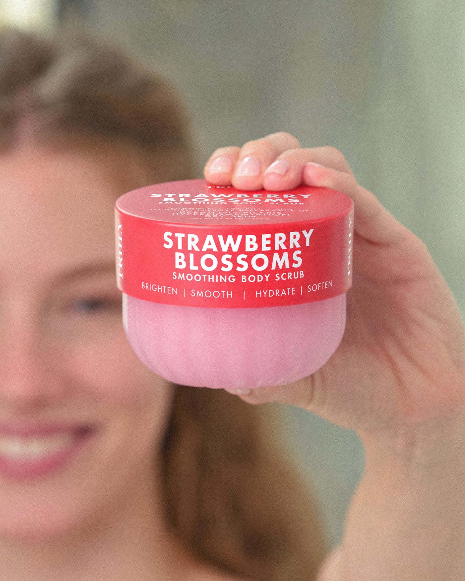 A person holding a jar of Strawberry Blossoms smoothing body scrub in front of their face, with the pink product jar and red lid clearly visible.