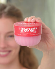 A person holding a jar of Strawberry Blossoms smoothing body scrub in front of their face, with the pink product jar and red lid clearly visible.