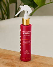A red spray bottle of Strawberry Blossoms body mist with a white trigger sprayer and gold collar, standing on a wooden surface with a blurred indoor background.
