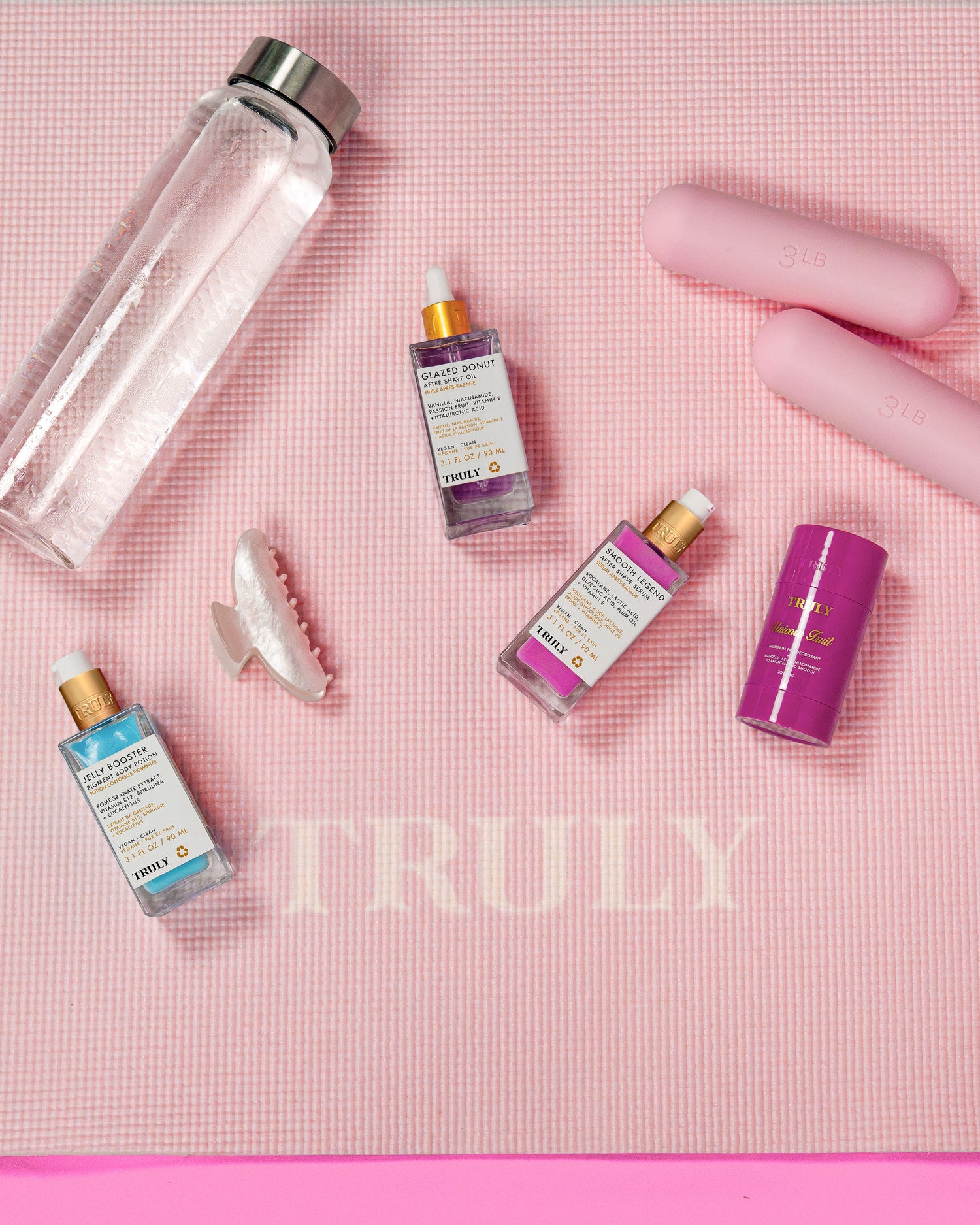 A pink exercise mat featuring “TRULY,” with a clear water bottle, two pink dumbbells, a white hair clip, a purple deodorant stick, and Truly Beauty’s Body Brightening Set for an even skin tone.