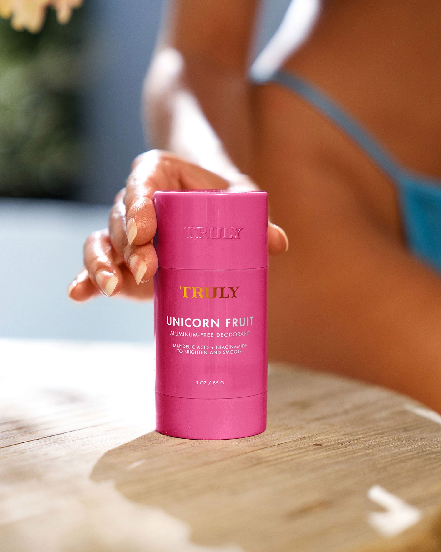 A person with manicured nails holds a pink stick of Truly Beauty Unicorn Fruit Skin Brightening and Smoothing Deodorant, sunlight highlighting the aluminum-free product against a softly blurred background.