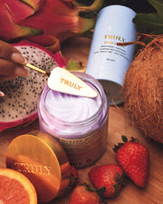 A hand holds a gold spatula labeled TRULY above a jar of Glazed Donut After Shave Glow Cream by Truly Beauty. Surrounded by strawberries, dragon fruit, coconut, and another skincare container, this cream delivers luscious hydration and glow.