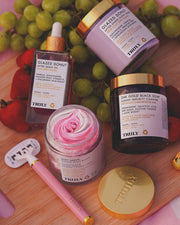 A flat lay features Glazed Donut After Shave Oil by Truly for post-shave hydration, a pink razor, 24K Gold Black Soap, Soft Serve Whipped Body Butter for soft skin, fresh grapes and strawberries, and a gold Truly lid on a wooden surface.