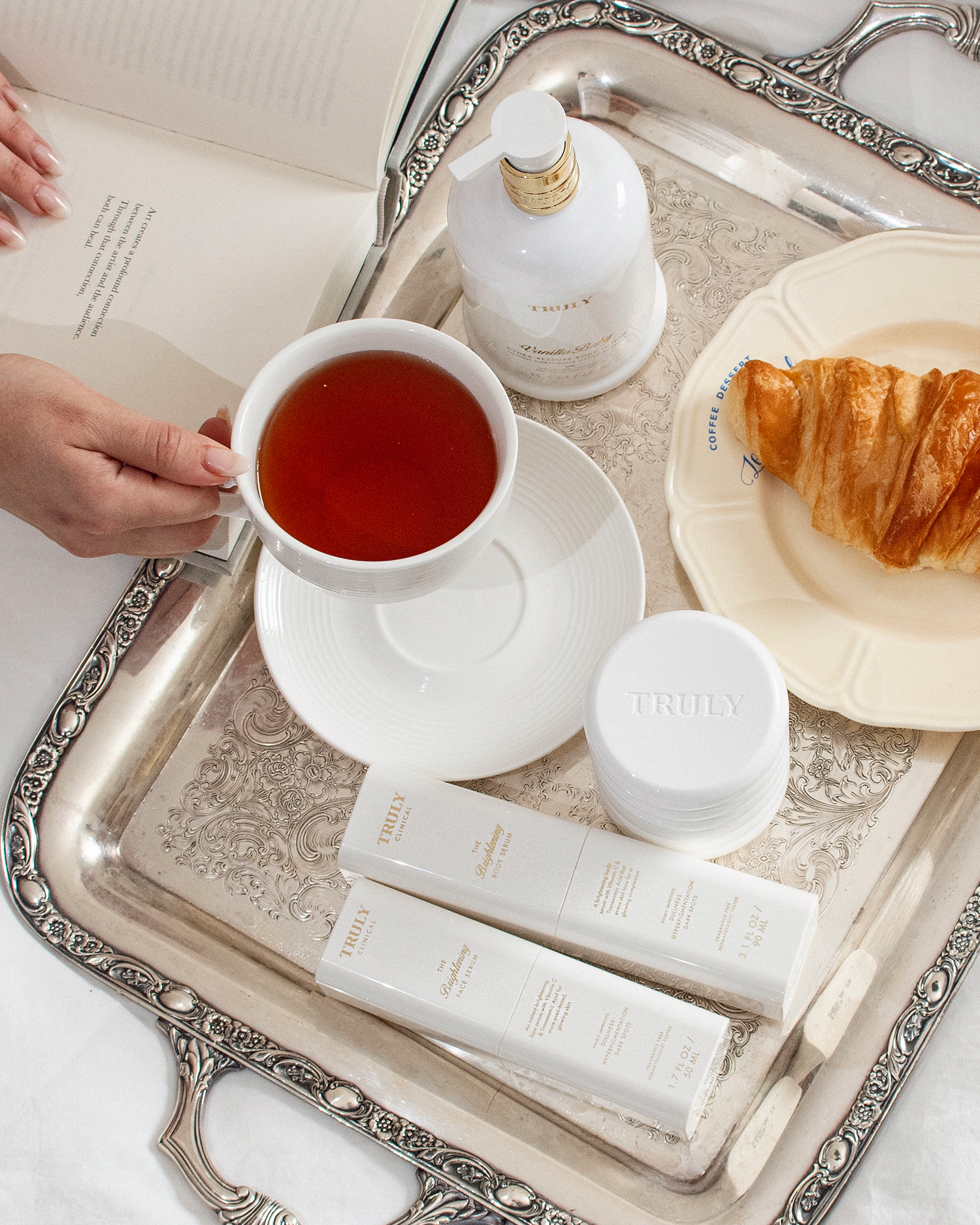 A hand holds a cup of tea on a silver tray with Truly Beauty skincare products, including the Cloud Cream Pro-Peptide Facial Moisturizer, a jar, two labeled tubes, a croissant on a plate, and a partly visible open book nearby.