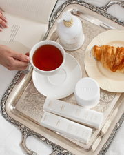 A hand holds a cup of tea on a silver tray with Truly Beauty skincare products, including the Cloud Cream Pro-Peptide Facial Moisturizer, a jar, two labeled tubes, a croissant on a plate, and a partly visible open book nearby.