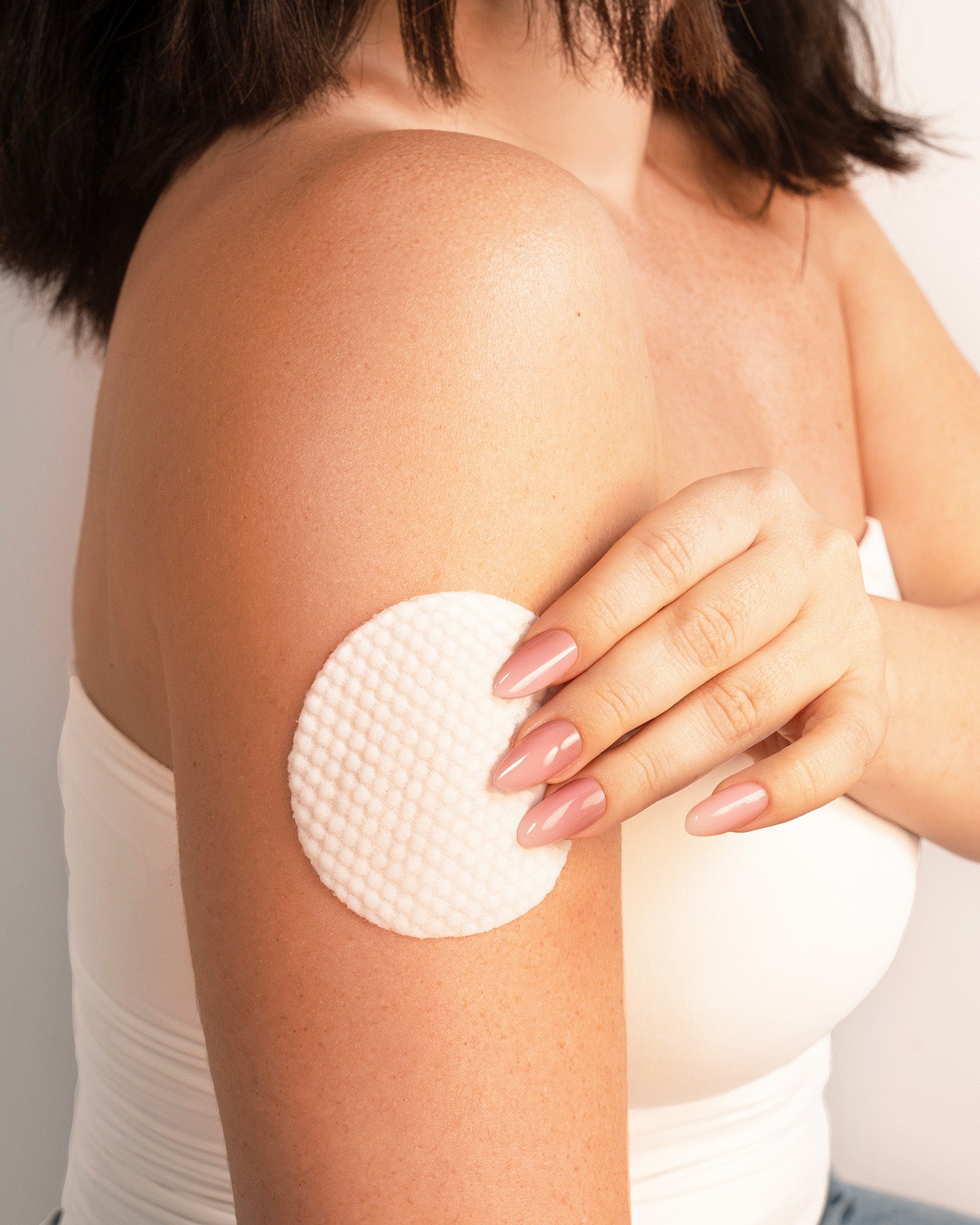 A woman wearing a white top holds up the Truly Beauty Strawberry Blossom Exfoliating & Resurfacing Face & Body Pad against her upper arm, her long nails painted a light pink shade.