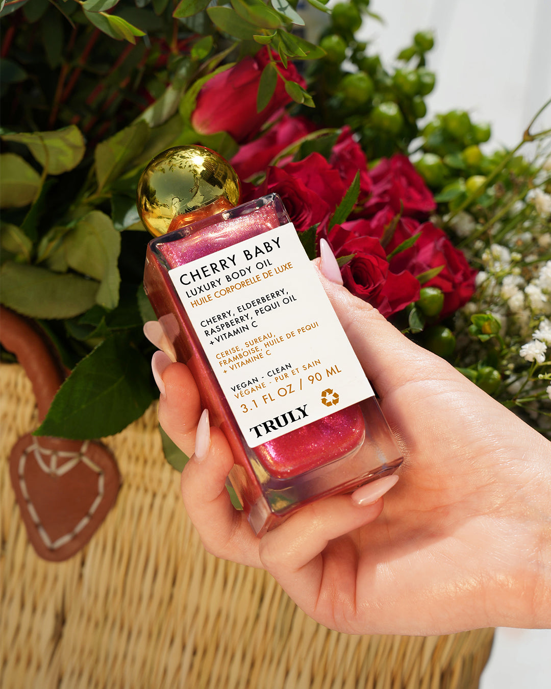 A manicured hand holds a bottle of Truly Beauty Cherry Baby Luxury Body Oil with a gold cap. The pink, shimmering oil promises radiant skin, while a wicker basket and a bouquet of red flowers enhance the elegant setting.