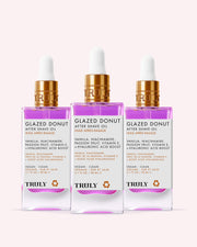 Three bottles of Truly Beauty’s Glazed Donut After Shave Oil (Copy), each with a gold dropper cap and clear label, are set against a light pink background—ideal for post-shave hydration and preventing ingrown hairs.