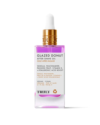 A pink and clear glass dropper bottle labeled Glazed Donut After Shave Oil, highlighting skin-smoothing niacinamide, vitamin E, and hyaluronic acid, is shown on a light pink background.