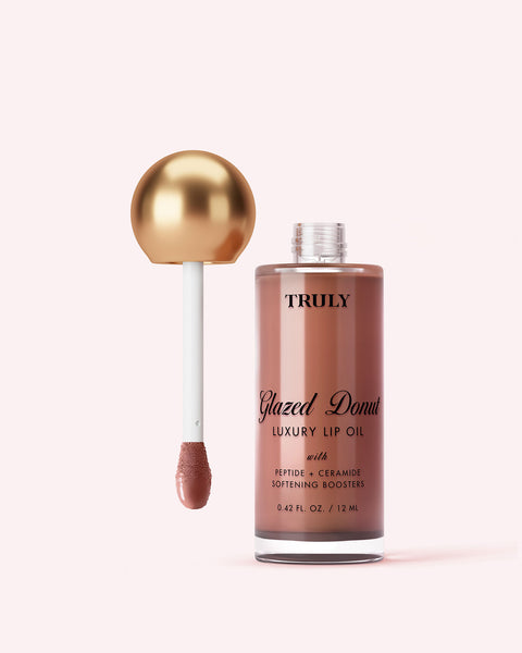 A bottle of Truly Beauty's Luxury Peptide Lip Oil with the applicator wand removed, revealing a gold sphere handle and pink-tinted oil, is set against a soft pink background.